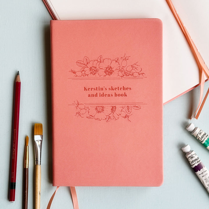 A5 vegan leather lined pink notebook personalised with the text Kerstin's sketches and ideas book, sits on a pale blur table surrounded by pencils, paintbrushes, and watercolour paint.