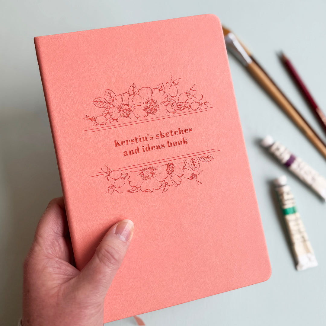 A pink journal personalised with the text Kerstin's sketches and ideas book, is held by a hand. In the background there are pencils, paintbrushes, and watercolour paint.