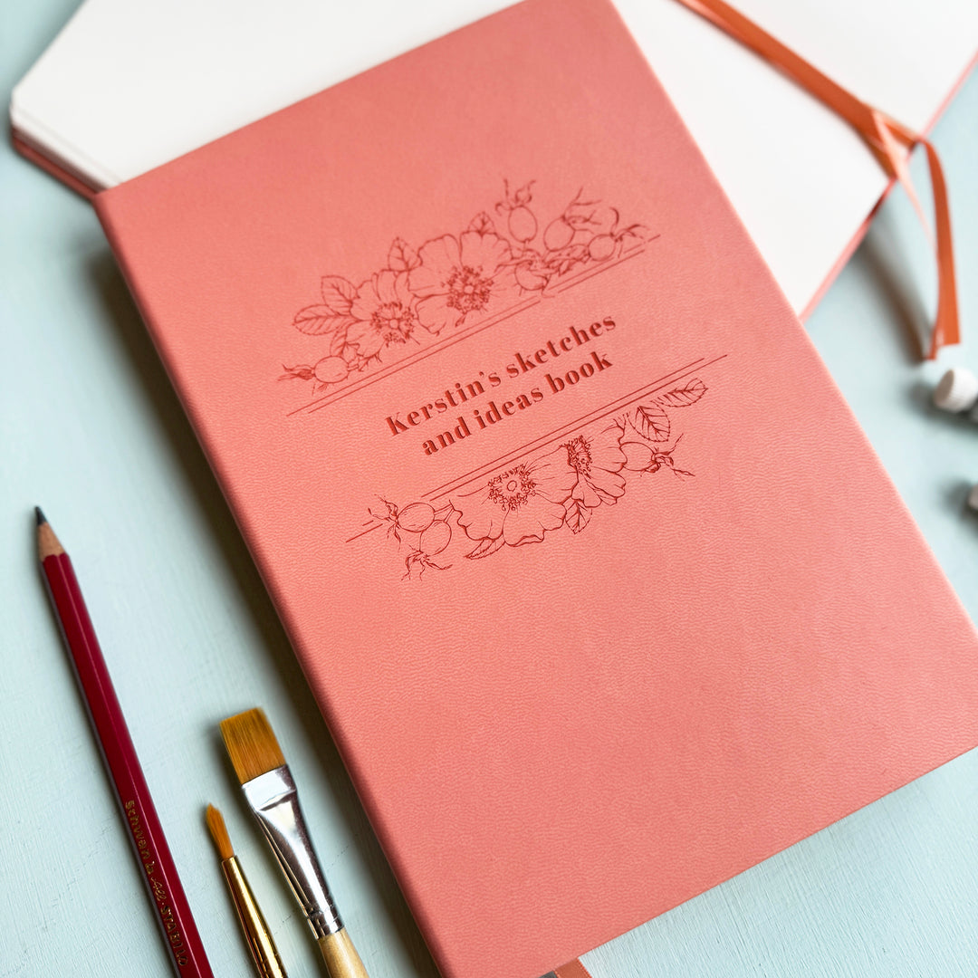 A pink notebook personalised with the text Kerstin's sketches and ideas book, sits on a pale blur table surrounded by pencils, paintbrushes, and watercolour paint.