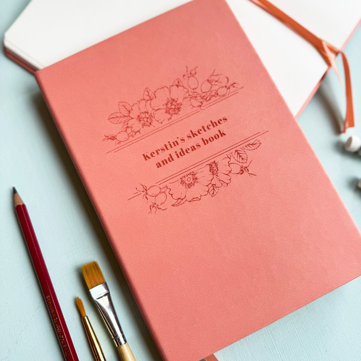 A pink notebook personalised with the text Kerstin's sketches and ideas book, sits on a pale blur table surrounded by pencils, paintbrushes, and watercolour paint.