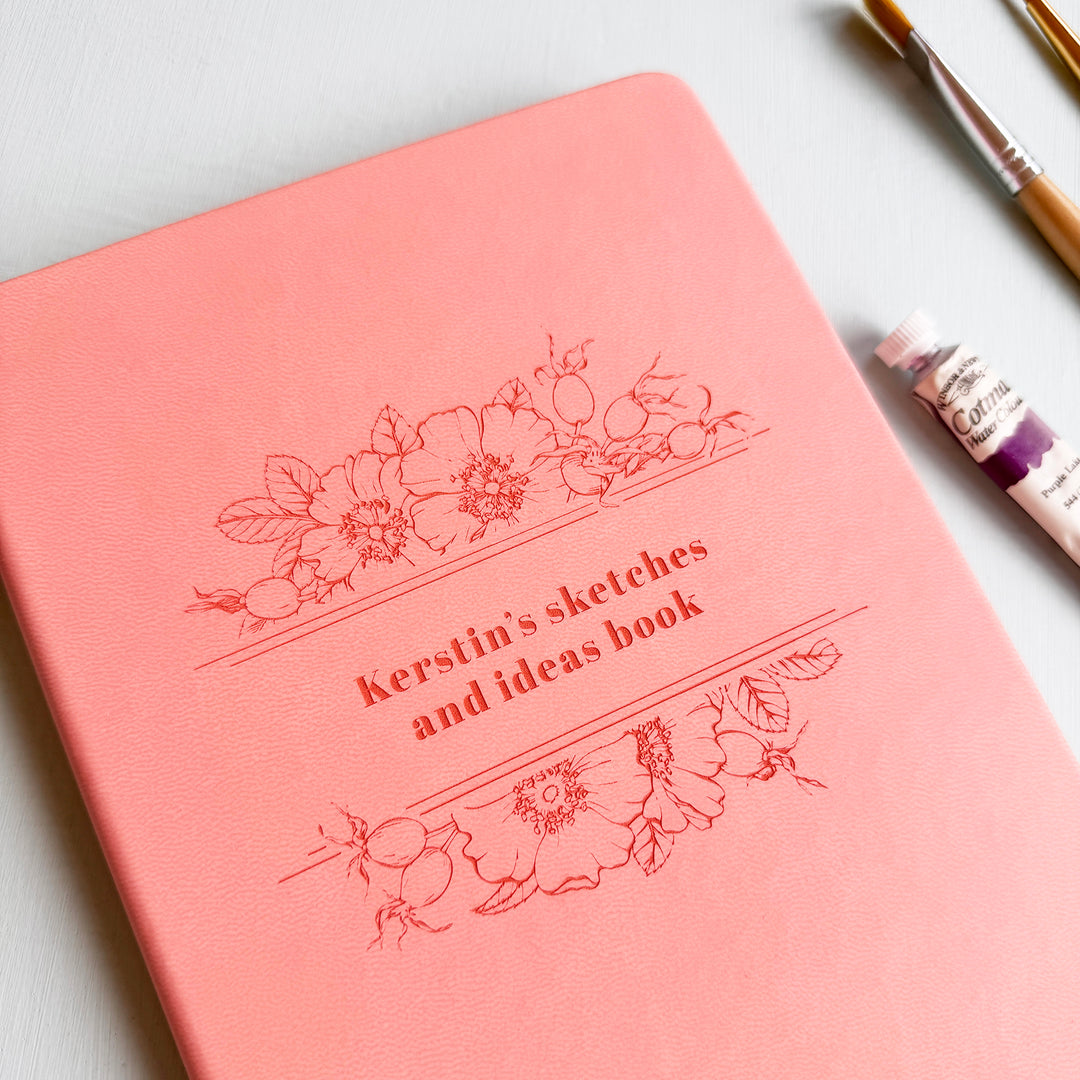 A pink notebook personalised with the text Kerstin's sketches and ideas book, sits on a pale blur table surrounded by pencils, paintbrushes, and watercolour paint.