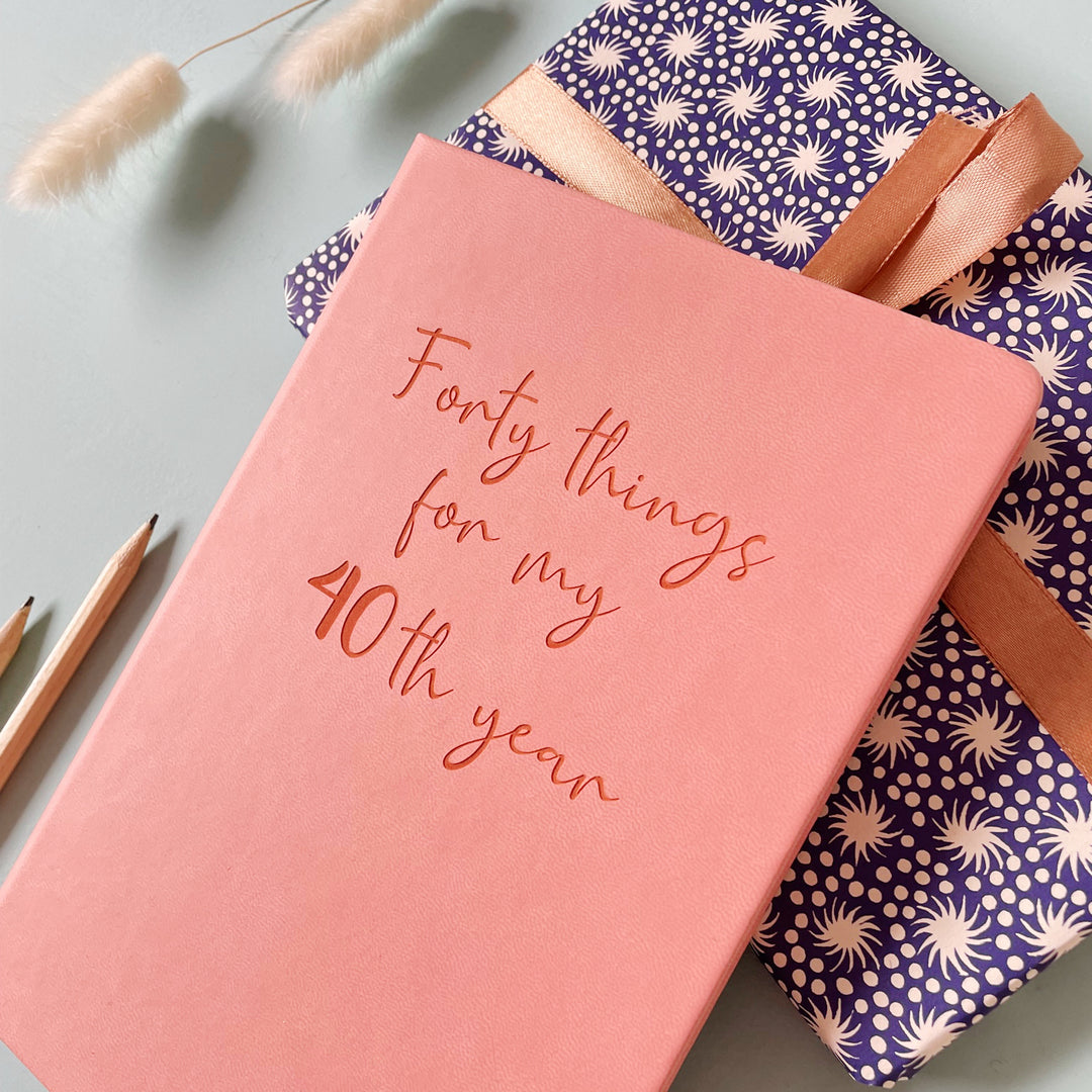 A top down close up image of a pink vegan leather notebook resting on a wrapped parcel with a ribbon. The front of the notebook features the words forty things for my 40th year. Surrounding the book are various stationary items and dried flowers.