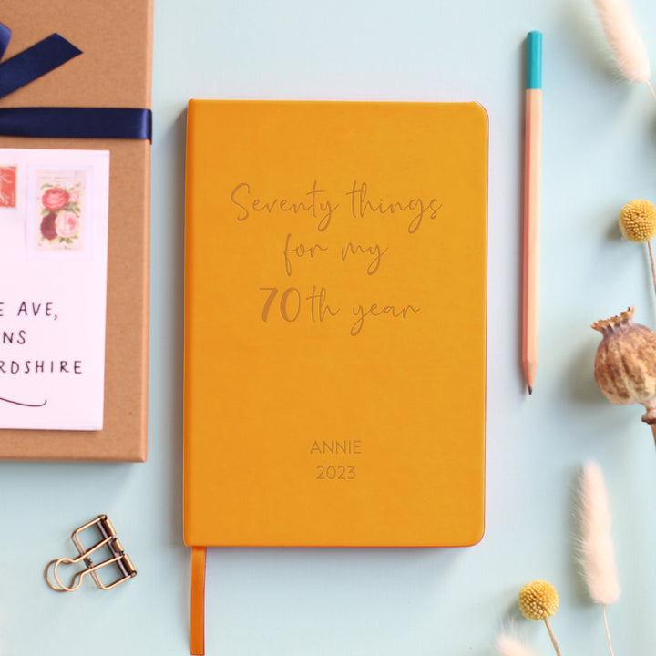 A top down image of a mustard yellow vegan leather notebook resting on a pale aqua table. The front of the notebook features the words Seventy things for my 70th year. The personalised engraving reads Annie 2023. Surrounding the book are various stationary items and dried flowers.