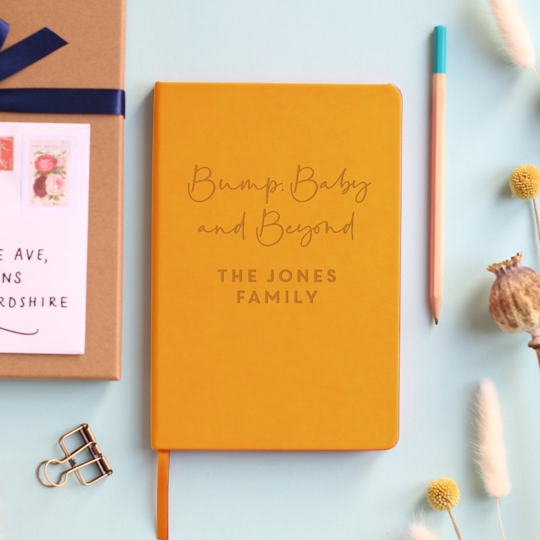 A top down image of a mustard yellow vegan leather notebook resting on a pale aqua table. The front of the notebook features the words Bump, Baby and Beyond. The personalised engraving reads The Jones Family. Surrounding the book are various stationary items and dried flowers.