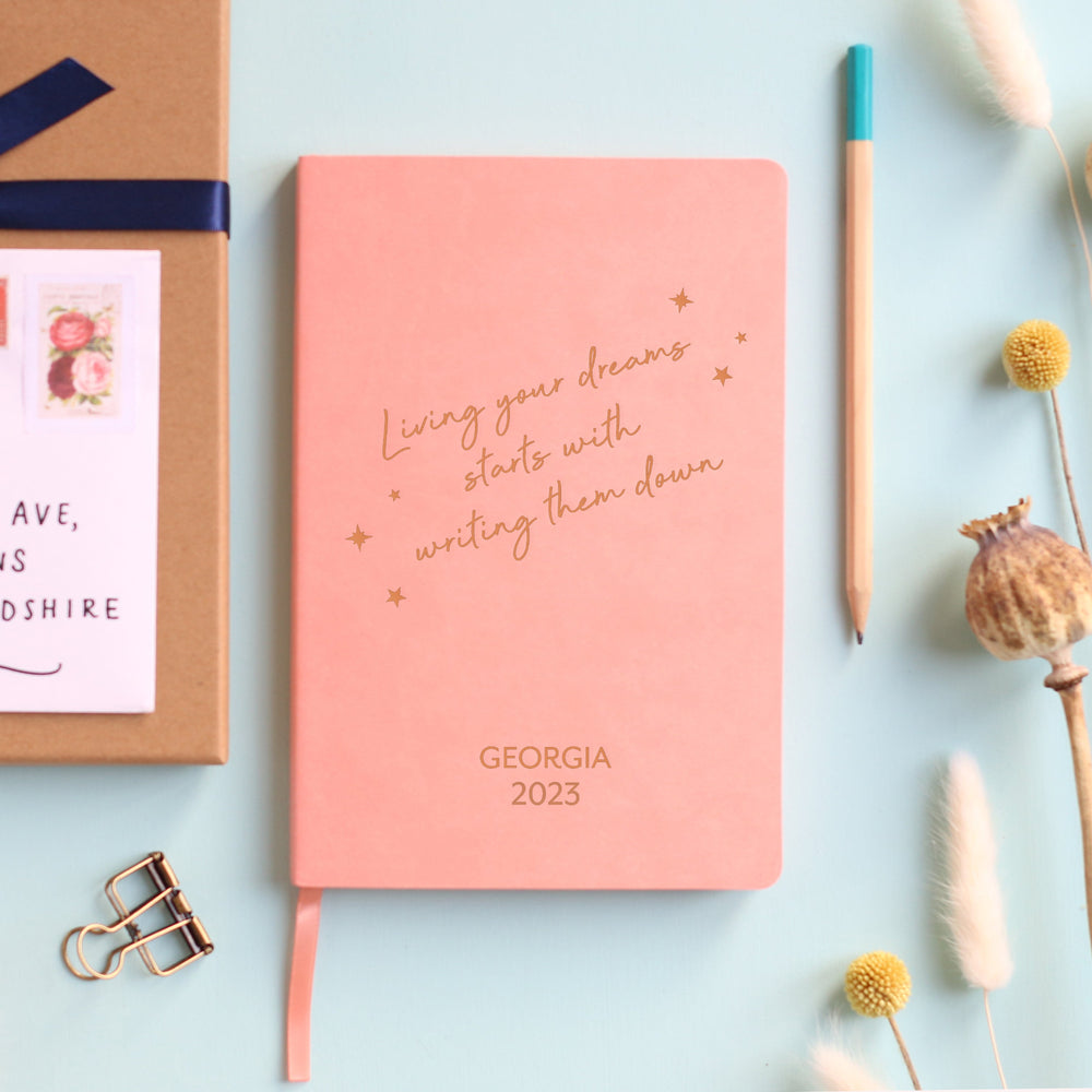 A top down image of a pink vegan leather notebook resting on a pale aqua table. The front of the notebook has the phrase “Living your dreams starts with writing them down” surrounded by stars. It is personalised with the text Georgia 2023. Surrounding the book are various stationary items and dried flowers.