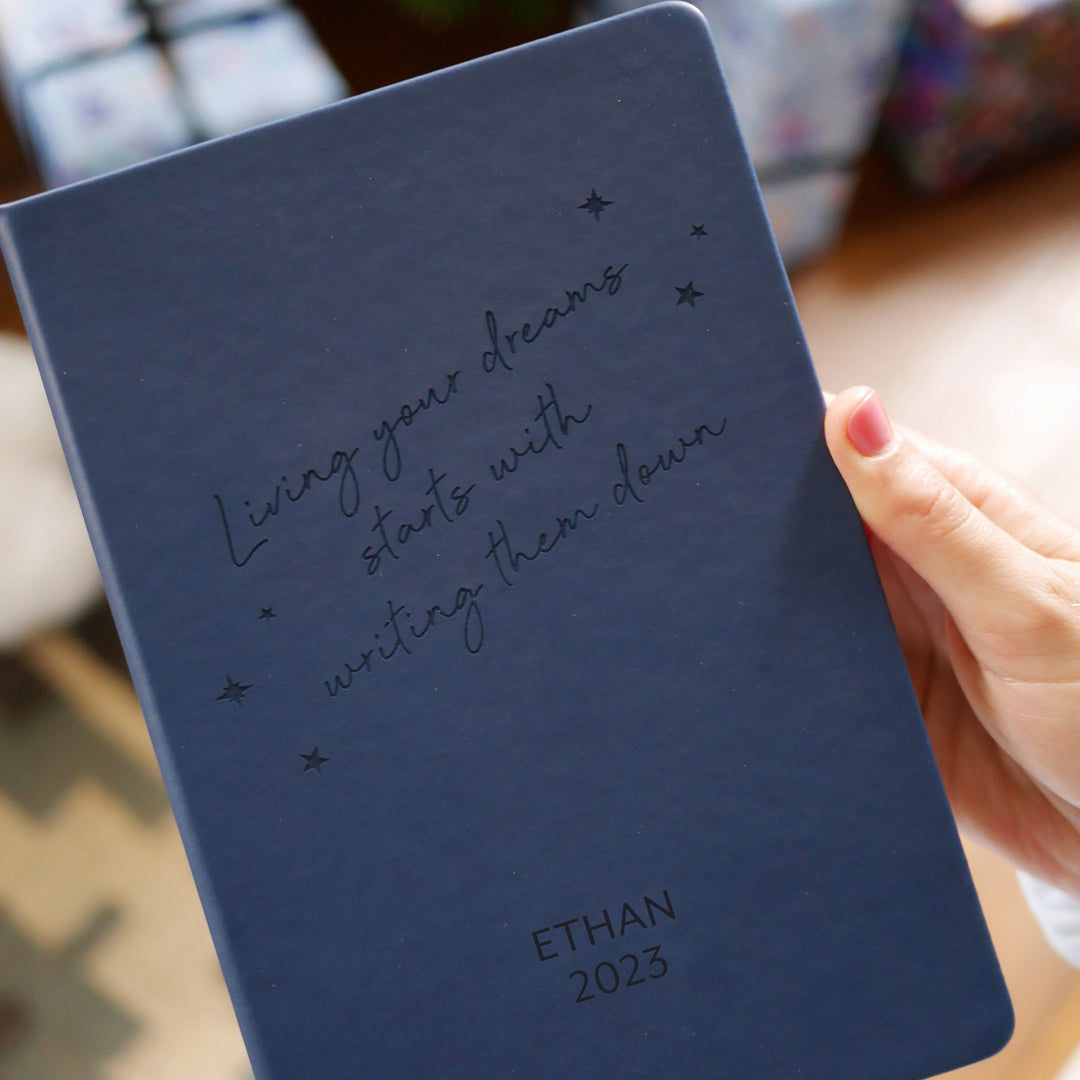 A close up image of woman's hands holding a navy blue vegan leather notebook. The front of the notebook has the phrase “Living your dreams starts with writing them down” surrounded by stars. It is personalised with the text Ethan 2023