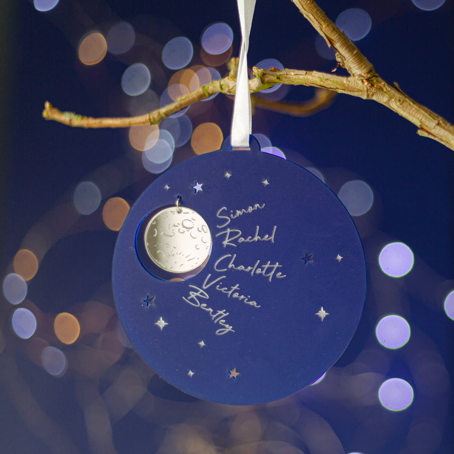 A personalized round Christmas tree decoration with a night sky and stars pattern, featuring engraved names, hanging on a tree branch.