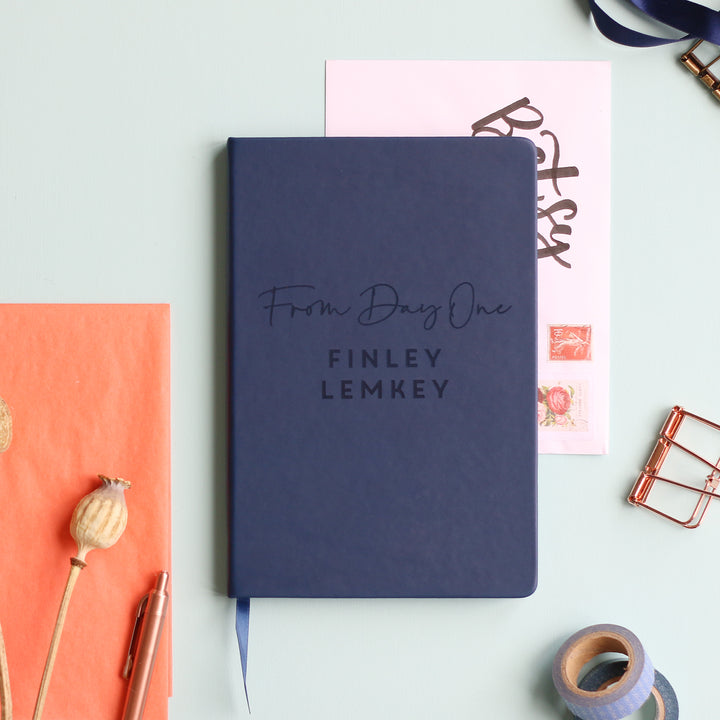 A top down image of a navy blue vegan leather notebook resting on a pale aqua table. The front of the notebook features the phrase "From Day One". It is personalised with the name Finley Lemkey. Surrounding the book are various stationary items and dried flowers.