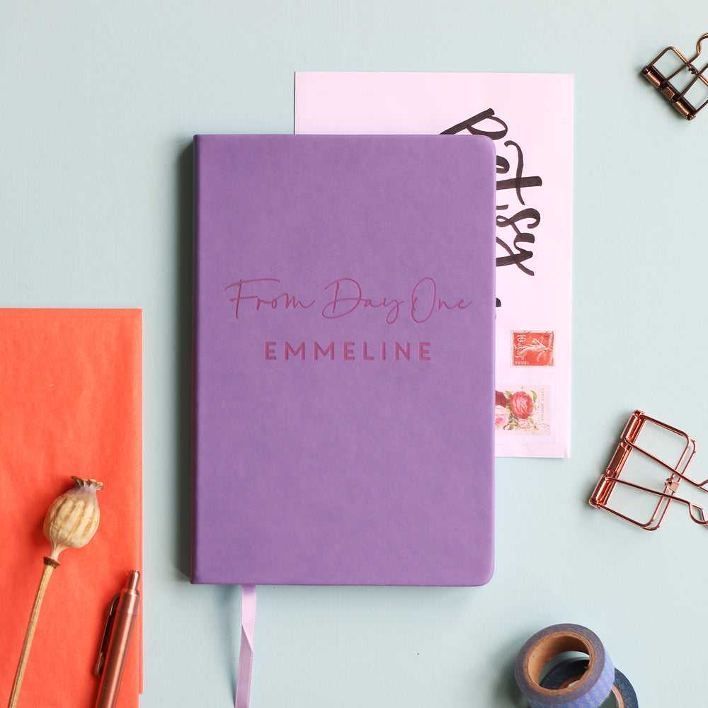 A top down image of a lilac vegan leather notebook resting on a pale aqua table. The front of the notebook features the phrase "From Day One". It is personalised with the name Emmeline. Surrounding the book are various stationary items and dried flowers.