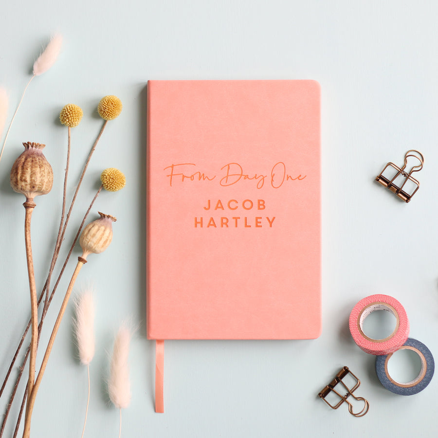 A top down image of a pink vegan leather notebook resting on a pale aqua table. The front of the notebook features the phrase "From Day One". It is personalised with the name Jacob Hartley. Surrounding the book are various stationary items and dried flowers.