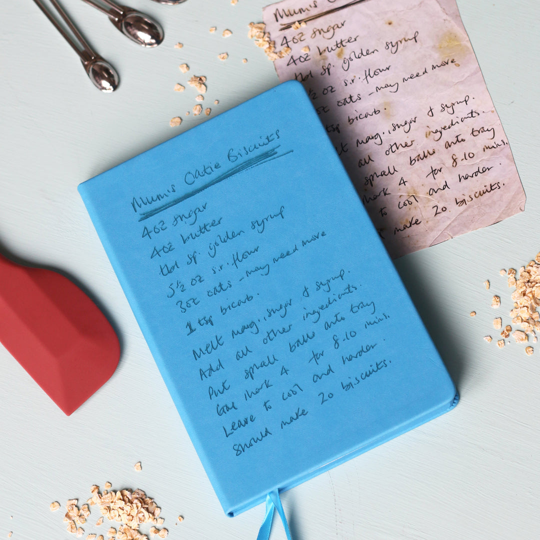 A top down image of a blue hardcover vegan leather notebook resting on a pale aqua table. The book is engraved with a handwritten recipe and the original paper recipe is lying next to the book. Surrounding the book are various kitchen utensils.