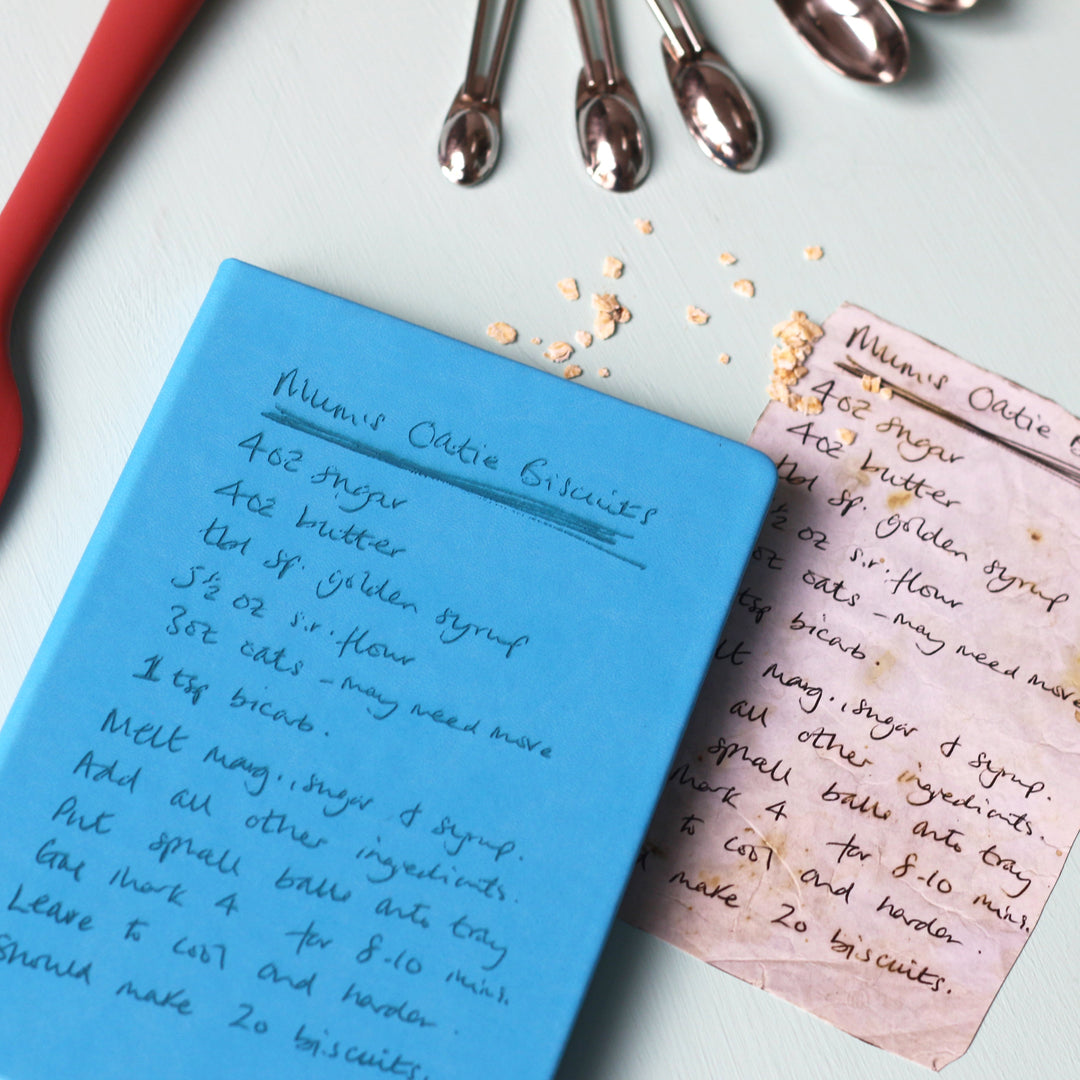 A top down image of a blue hardcover vegan leather notebook resting on a pale aqua table. The book is engraved with a handwritten recipe and the original paper recipe is lying next to the book. Surrounding the book are various kitchen utensils.