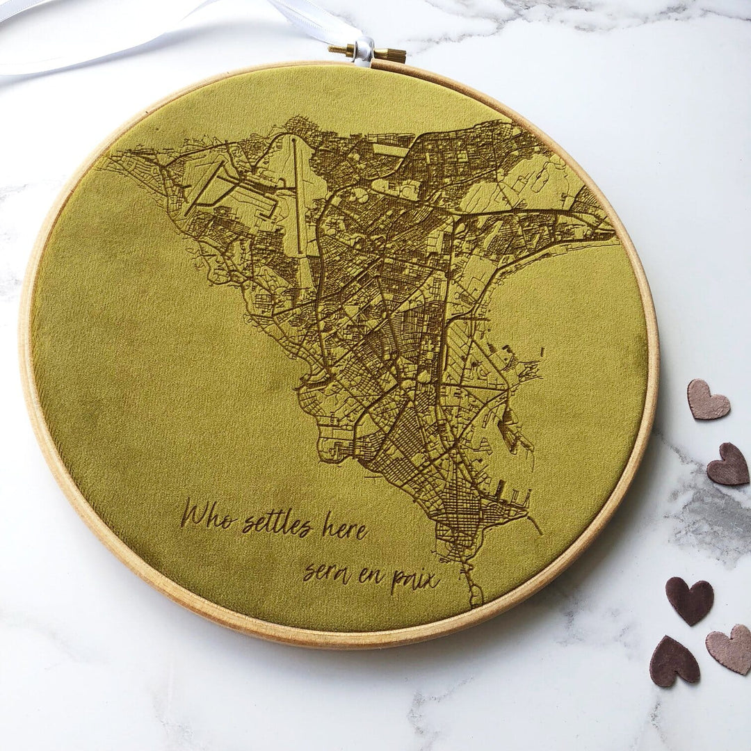 A circular embroidery hoop with a green velvet map of a city embroidered on it. The text "Who settles here, sera en paix" is embroidered at the bottom. The hoop is surrounded by small decorative hearts.