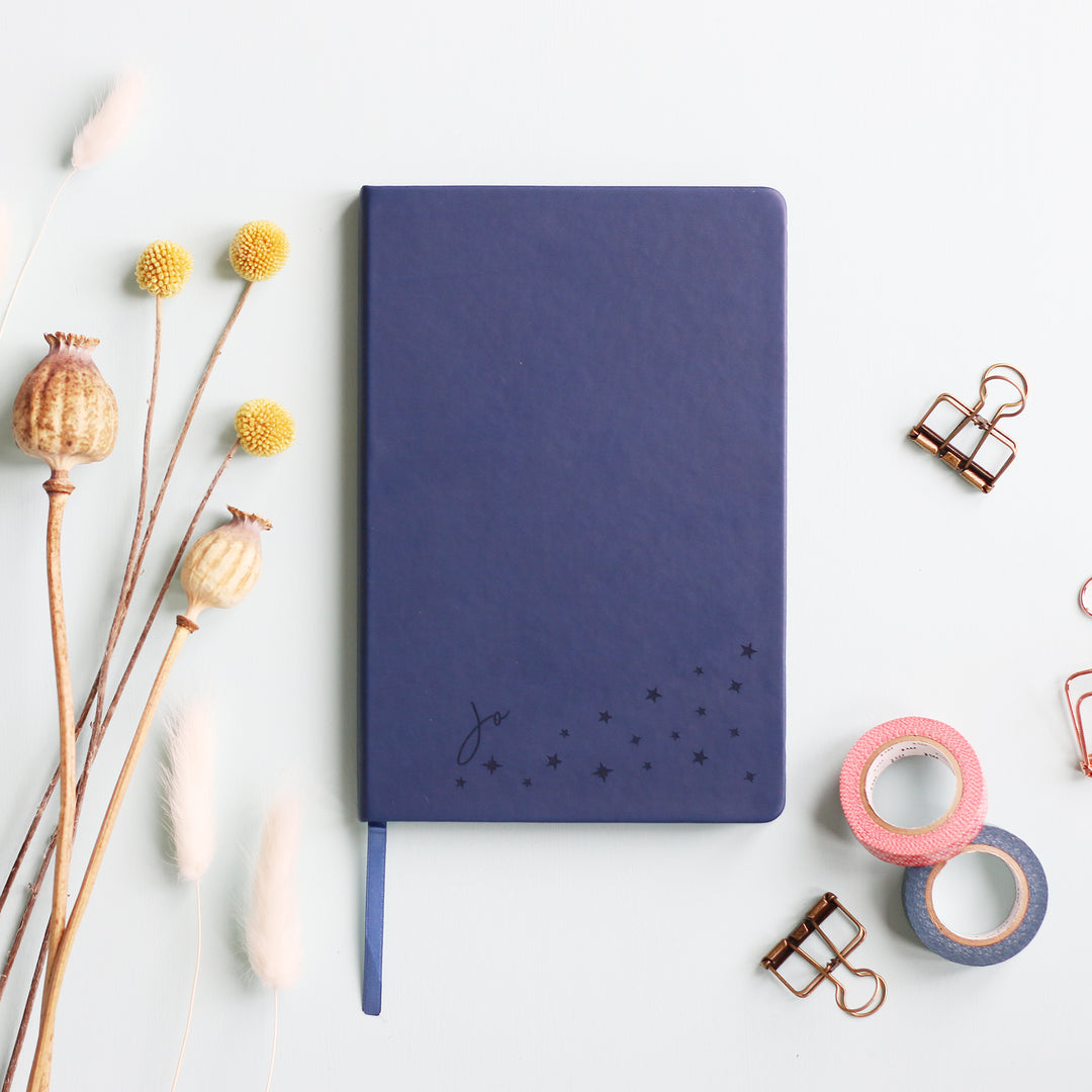 A flat lay image of a navy blue vegan leather notebook resting on a pale aqua table. The front of the notebook is personalised with the name Jo above a cluster of different sized engraved stars. Surrounding the book are various stationary items and dried flowers.