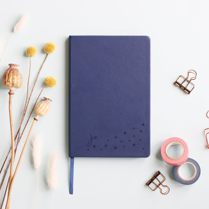 A flat lay image of a navy blue vegan leather notebook resting on a pale aqua table. The front of the notebook is personalised with the name Jo above a cluster of different sized engraved stars. Surrounding the book are various stationary items and dried flowers.