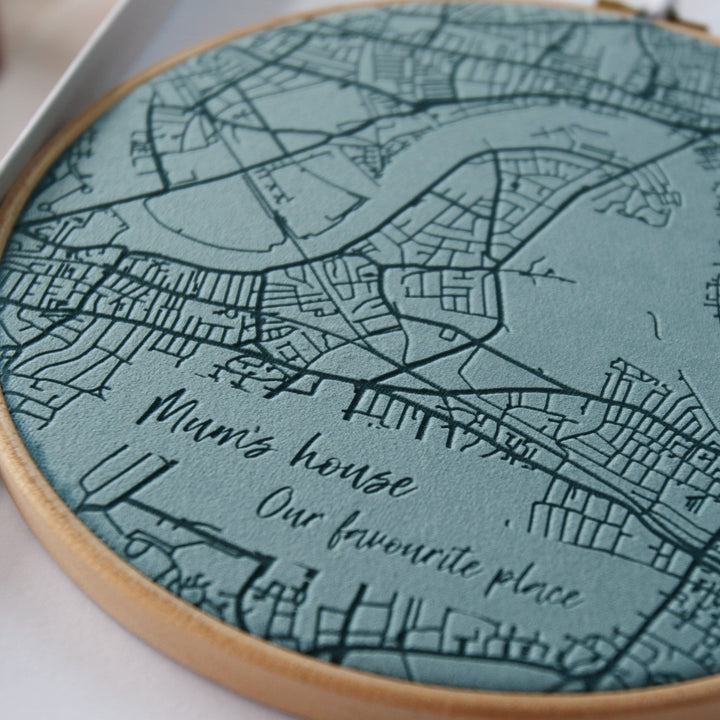 A close-up of a teal fabric map embroidered in a circular hoop. The map features roads, rivers, and the text "Mum's house. Our favourite place."