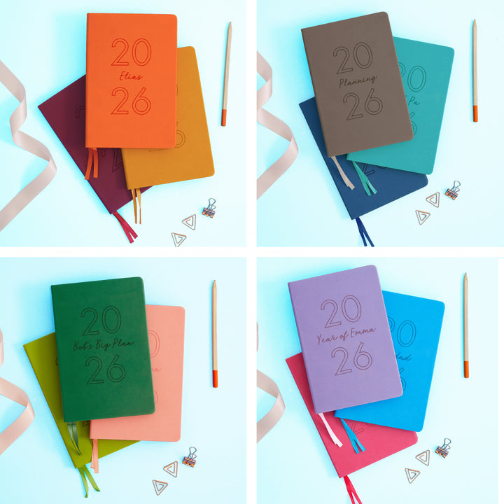 The image shows examples of personalised engraved text on all 12 colours of notebook. The books are stacked in in threes and are surround by various pieces of stationery.