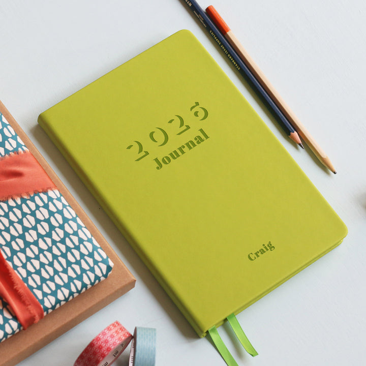 A top down image of a lime green vegan leather notebook resting on a pale aqua table. The front of the notebook features the words 2026 Journal. The personalised engraving reads Craig. Surrounding the book are various stationary items.