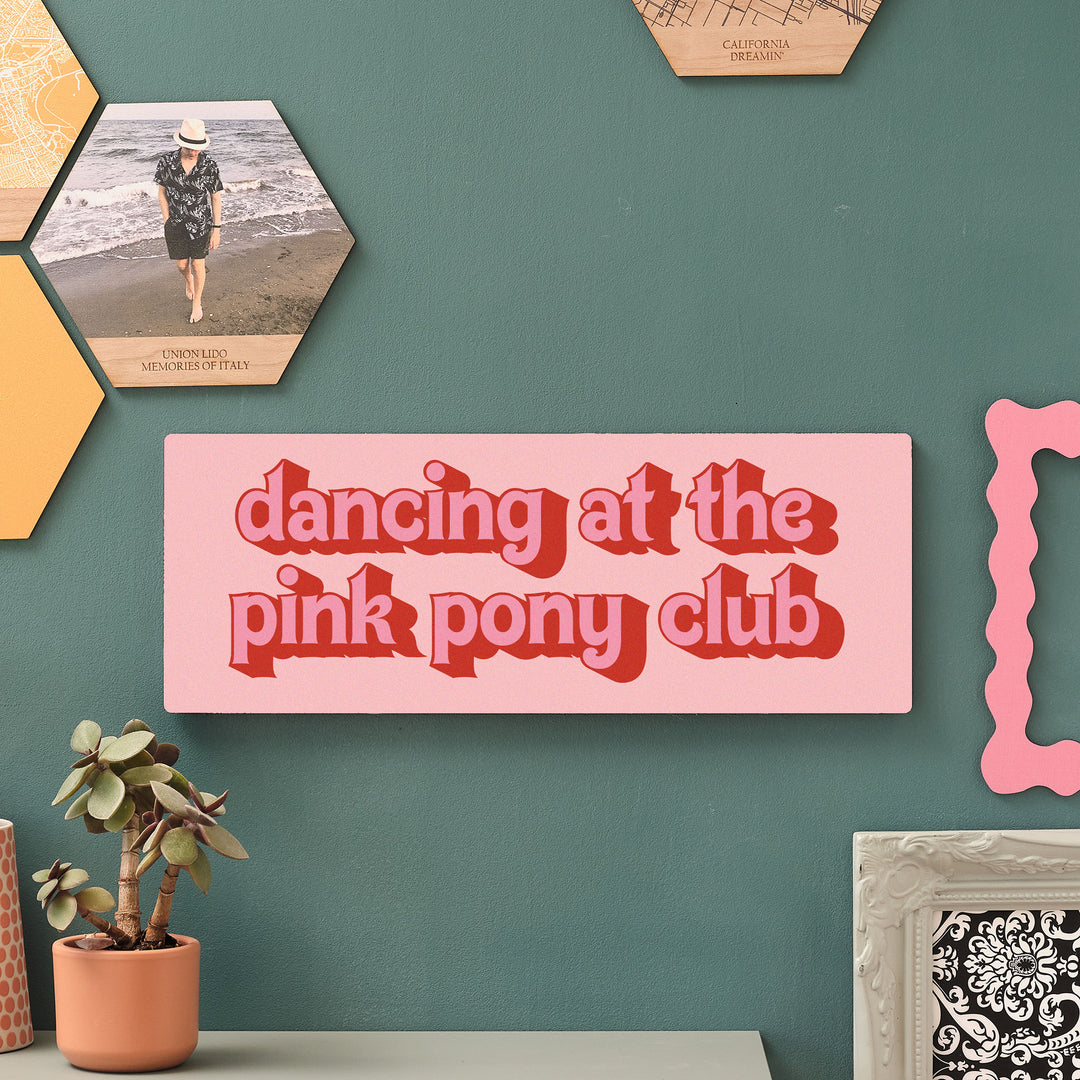 Decorative sign with 'dancing at the pink pony club' text on a teal wall with hexagon printed wooden pictures and a plant.