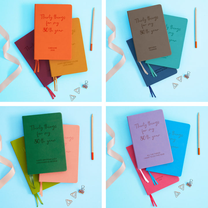 The image shows examples of personalised engraved text on all 12 colours of notebook. The books are stacked in threes and are surrounded by various pieces of stationery.
