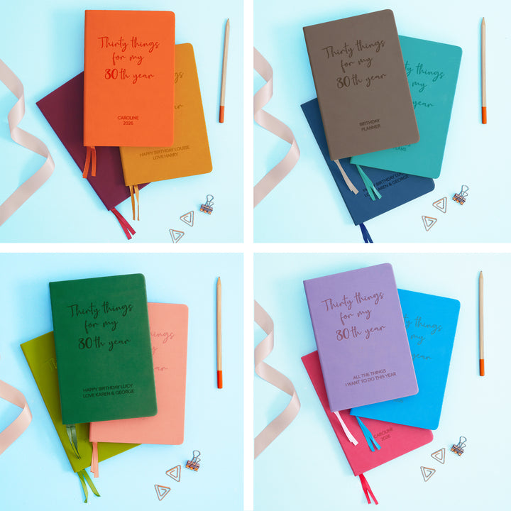 The image shows examples of personalised engraved text on all 12 colours of notebook. The books are stacked in in threes and are surround by various pieces of stationery.
