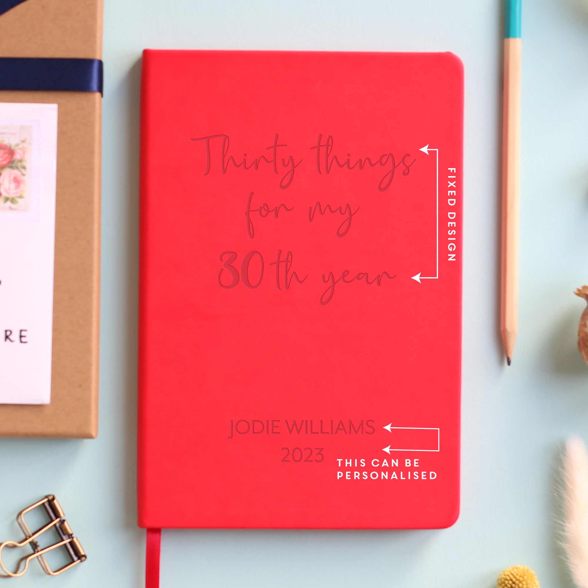 A top down image of a pink vegan leather notebook resting on a pale aqua table. The front of the notebook features the words thirty things for my 30th year. The personalised engraving reads Happy Birthday Love Tracey and Carl. Surrounding the book are various stationary items and dried flowers.