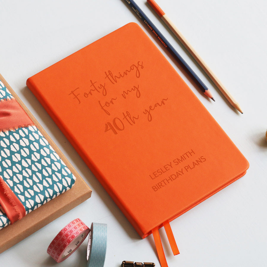A top down image of a orange vegan leather notebook resting on a pale aqua table. The front of the notebook features the words forty things for my 40th year. The personalised engraving reads Lesley Smith Birthday Plans. Surrounding the book are various stationary items.