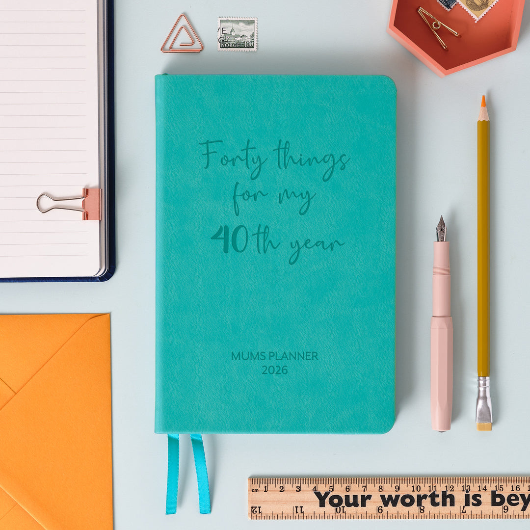 A top down image of a turquoise vegan leather notebook resting on a pale aqua table. The front of the notebook features the words forty things for my 40th year. The personalised engraving reads Mums Planner 2026. Surrounding the book are various stationary items and dried flowers.