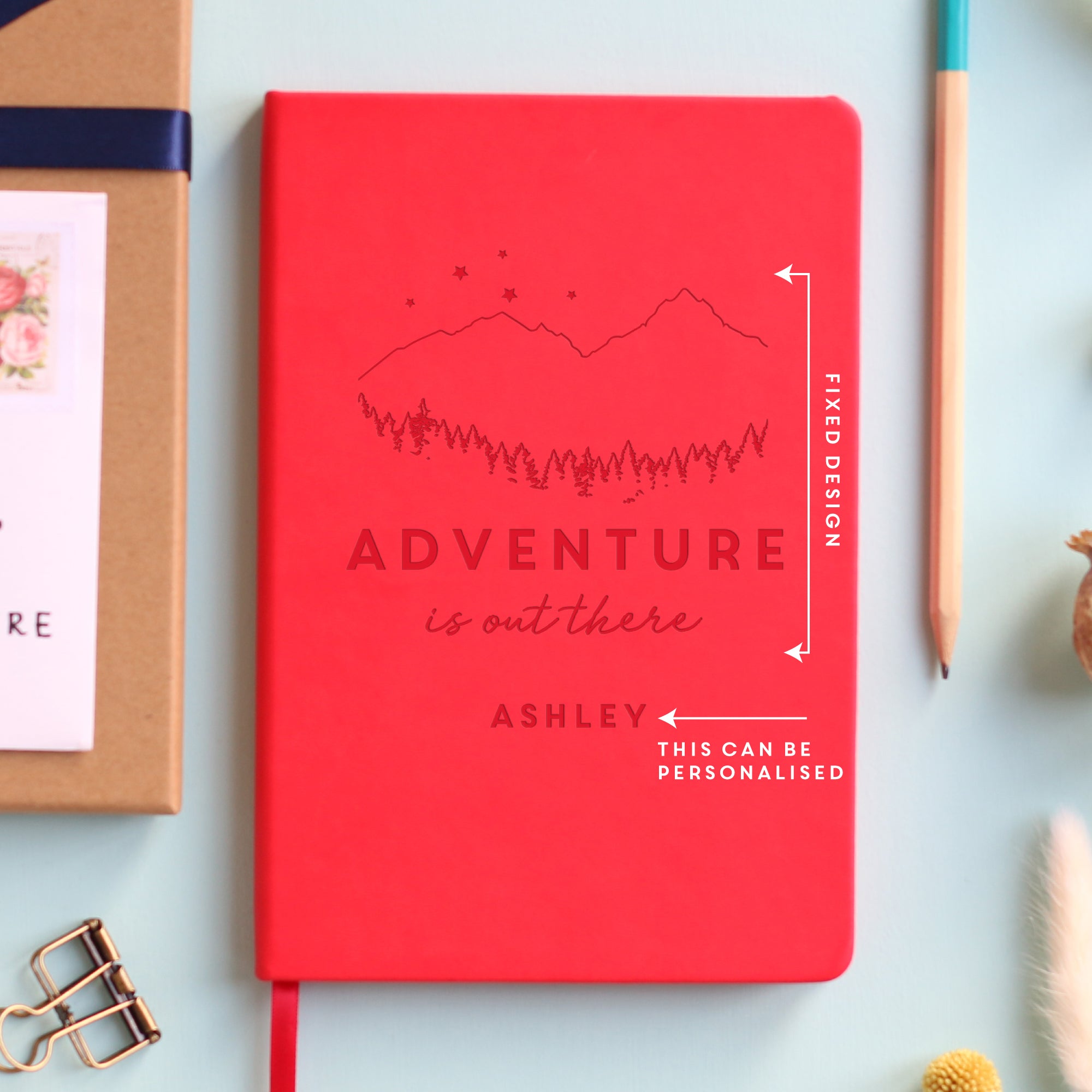 A top down image of a red vegan leather notebook resting on a pale aqua table. The front of the notebook features an engraving of a mountain scene with stars and the text Adventure is out there. Personalised with the name Ashley. Surrounding the book are various stationary items.