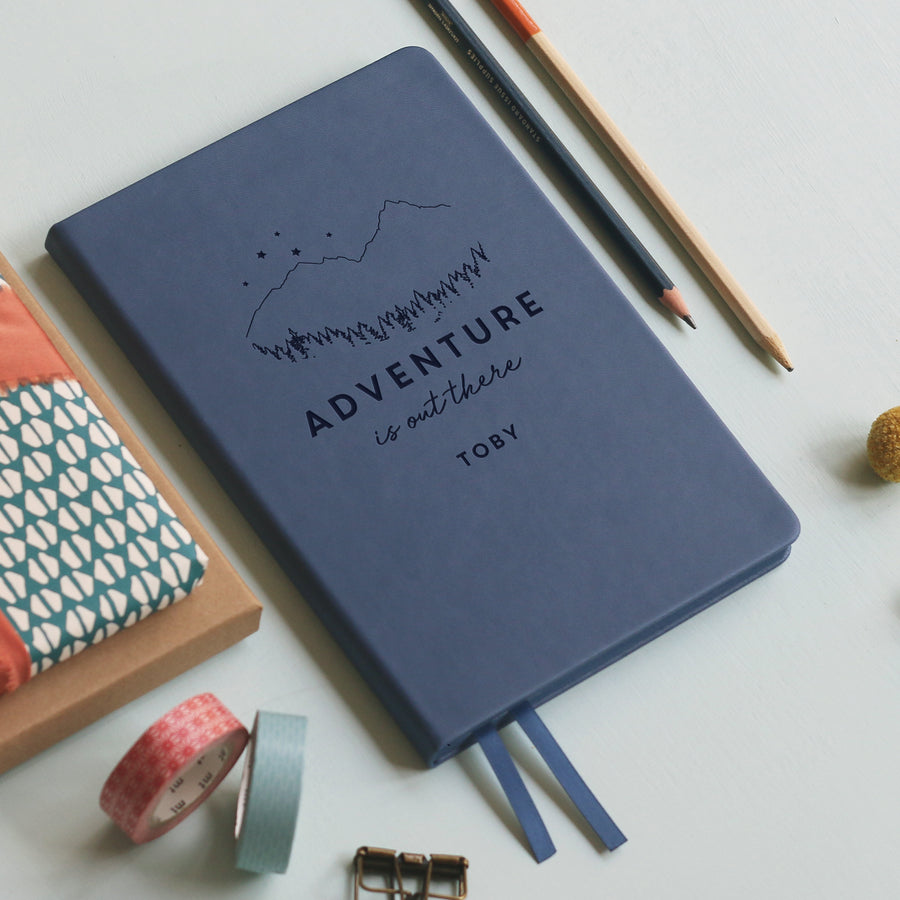 A flat lay image of a mid blue vegan leather notebook resting on a pale aqua table. The front of the notebook is is engraved with the phrase 'Adventure is out there' with the name Toby personalised below. Surrounding the book are various stationary items.