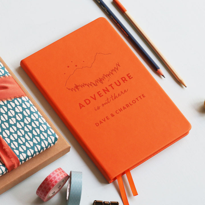 A flat lay image of a orange vegan leather notebook resting on a pale aqua table. The front of the notebook is is engraved with the phrase 'Adventure is out there' with Dave & Charlotte personalised below. Surrounding the book are various stationary items.