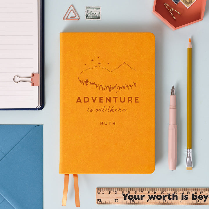 A flat lay image of a mustard yellow vegan leather notebook resting on a pale aqua table. The front of the notebook is is engraved with the phrase 'Adventure is out there' with the name Ruth personalised below. Surrounding the book are various stationary items.