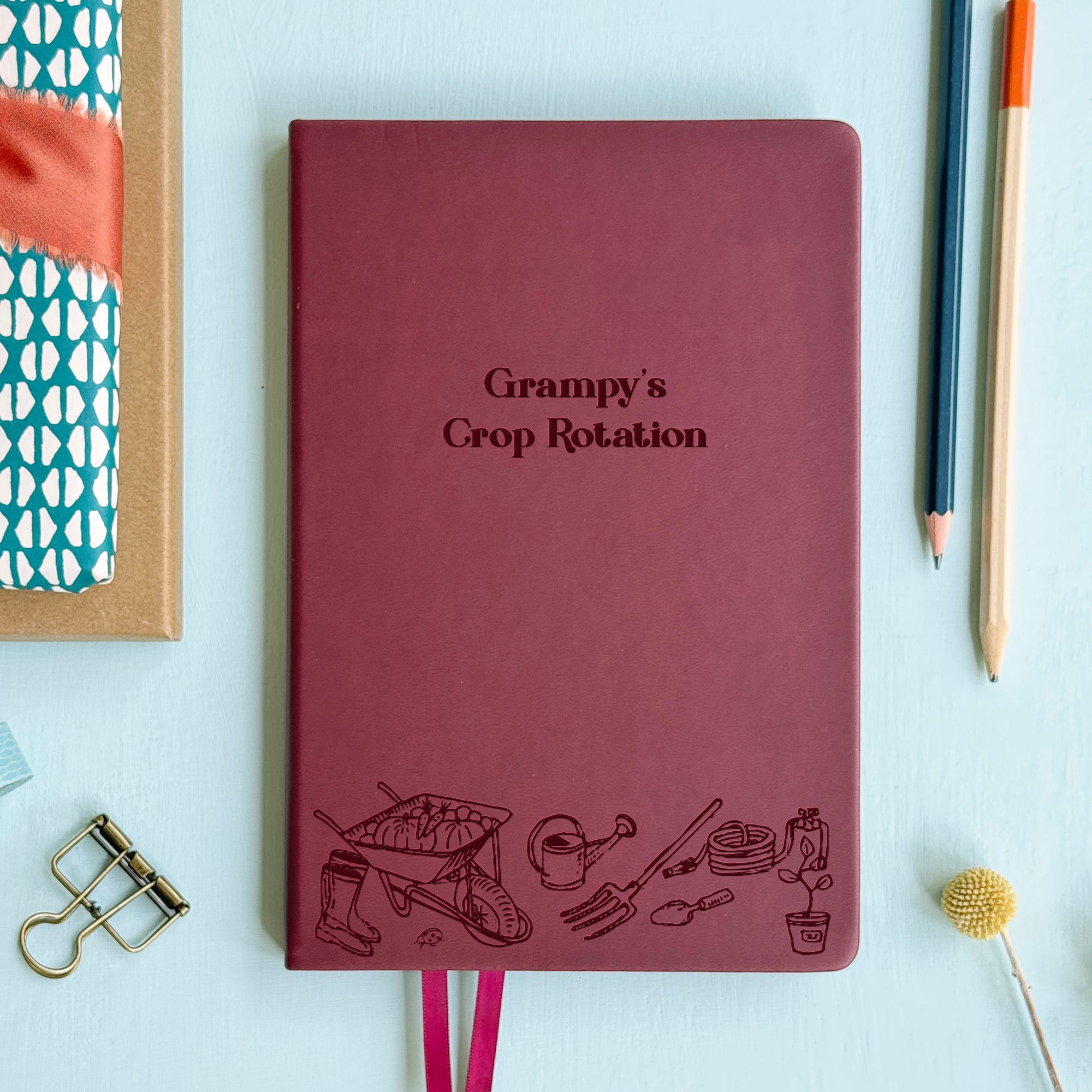 A top down image of a burgundy vegan leather notebook resting on a pale aqua table. The front of the notebook features an engraving of gardening tools along the bottom edge. The personalised text reads 