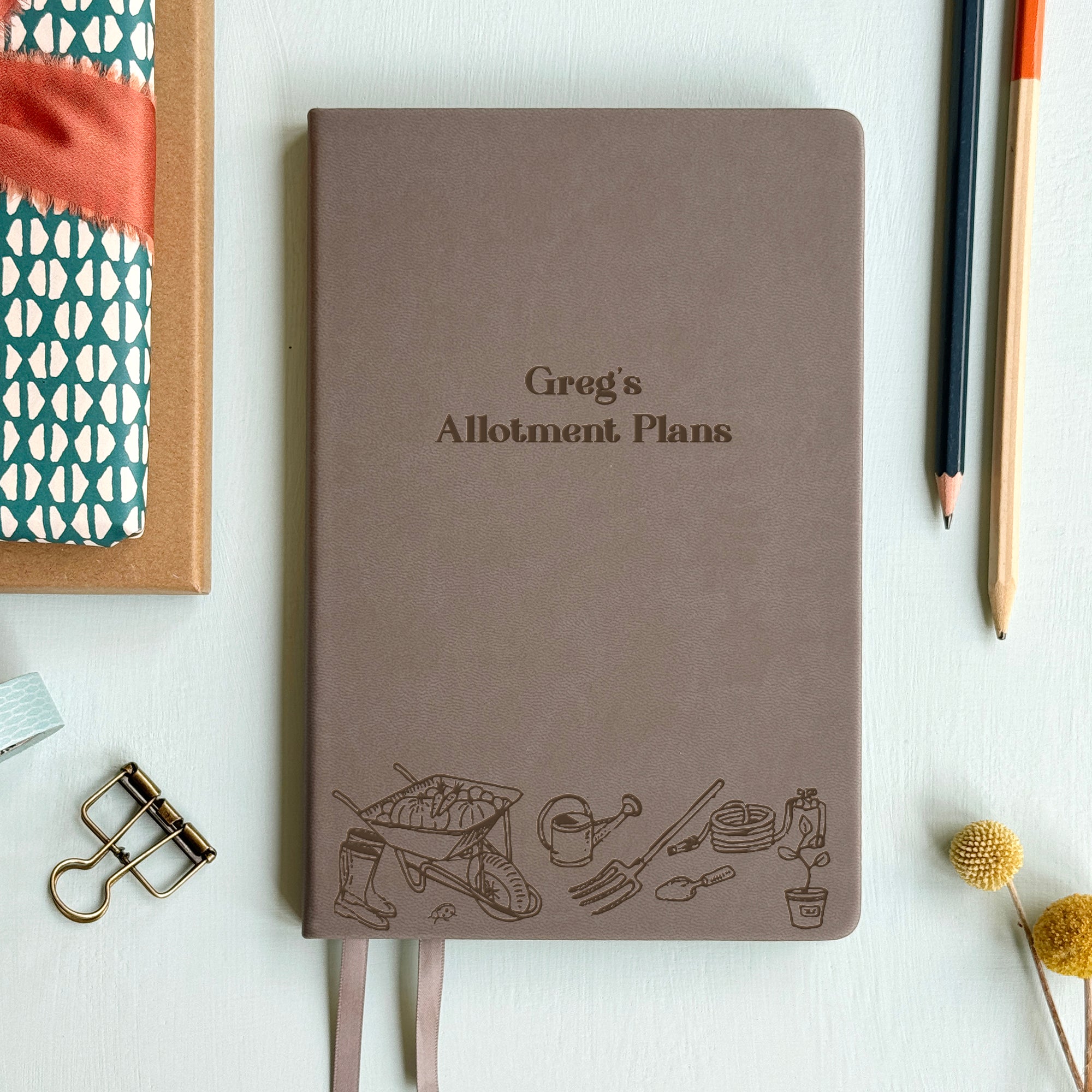 A top down image of a grey vegan leather notebook resting on a pale aqua table. The front of the notebook features an engraving of gardening tools along the bottom edge. The personalised text reads Greg's Allotment Plans. Surrounding the book are various stationary items and dried flowers.