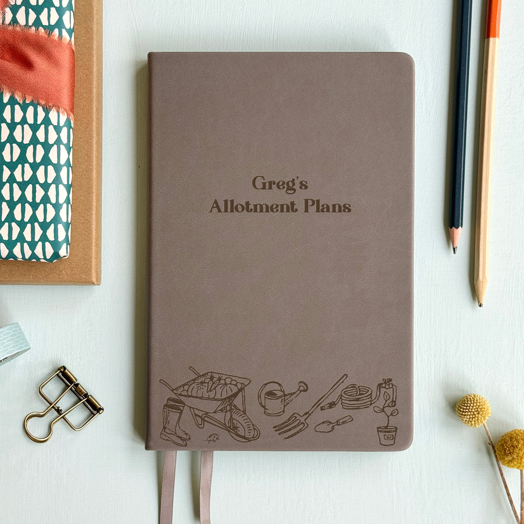 A top down image of a grey vegan leather notebook resting on a pale aqua table. The front of the notebook features an engraving of gardening tools along the bottom edge. The personalised text reads Greg's Allotment Plans. Surrounding the book are various stationary items and dried flowers.