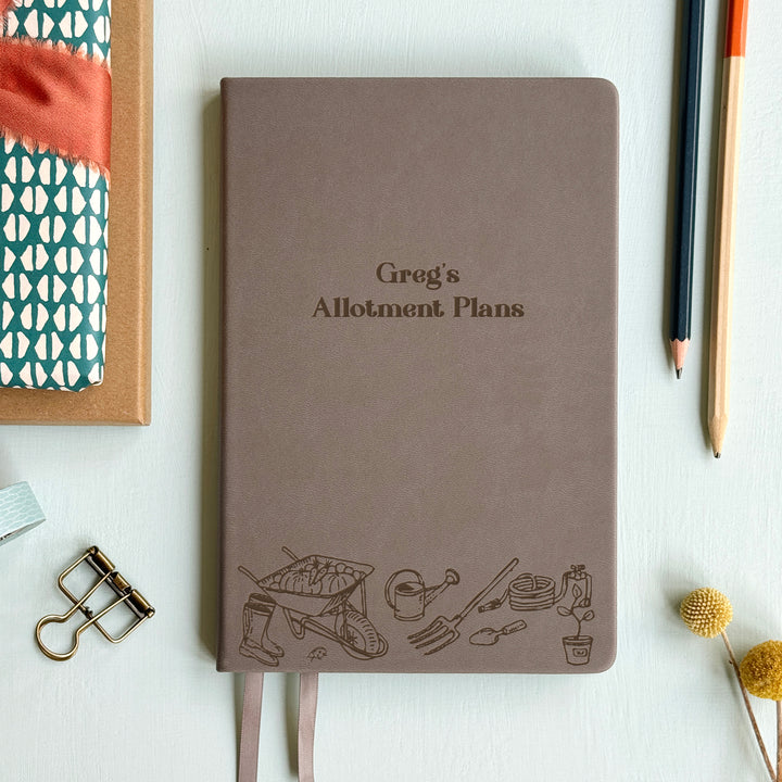 A top down image of a grey vegan leather notebook resting on a pale aqua table. The front of the notebook features an engraving of gardening tools along the bottom edge. The personalised text reads Greg's Allotment Plans. Surrounding the book are various stationary items and dried flowers.