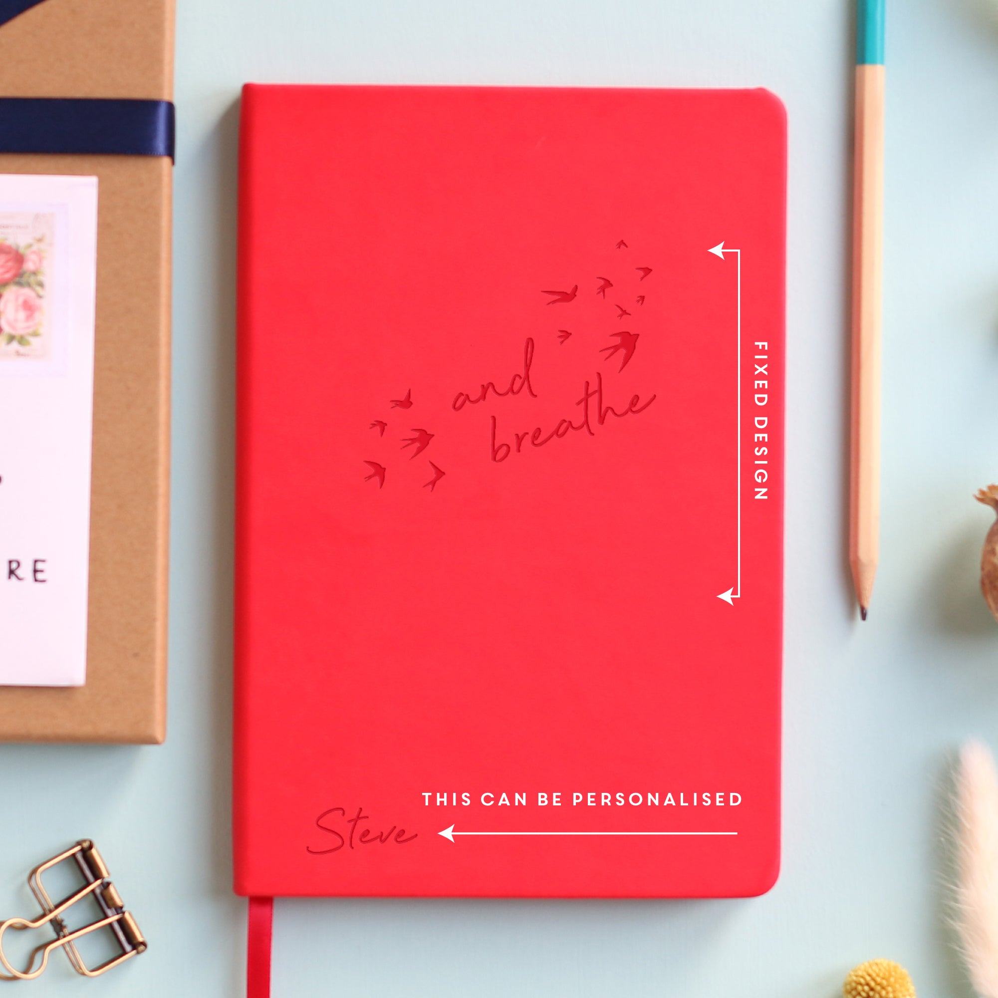 A top down image of a red vegan leather notebook resting on a pale aqua table. The front of the notebook features the words "and breathe" surrounded by birds. The personalised engraving reads Steve. Surrounding the book are various stationary items and dried flowers.