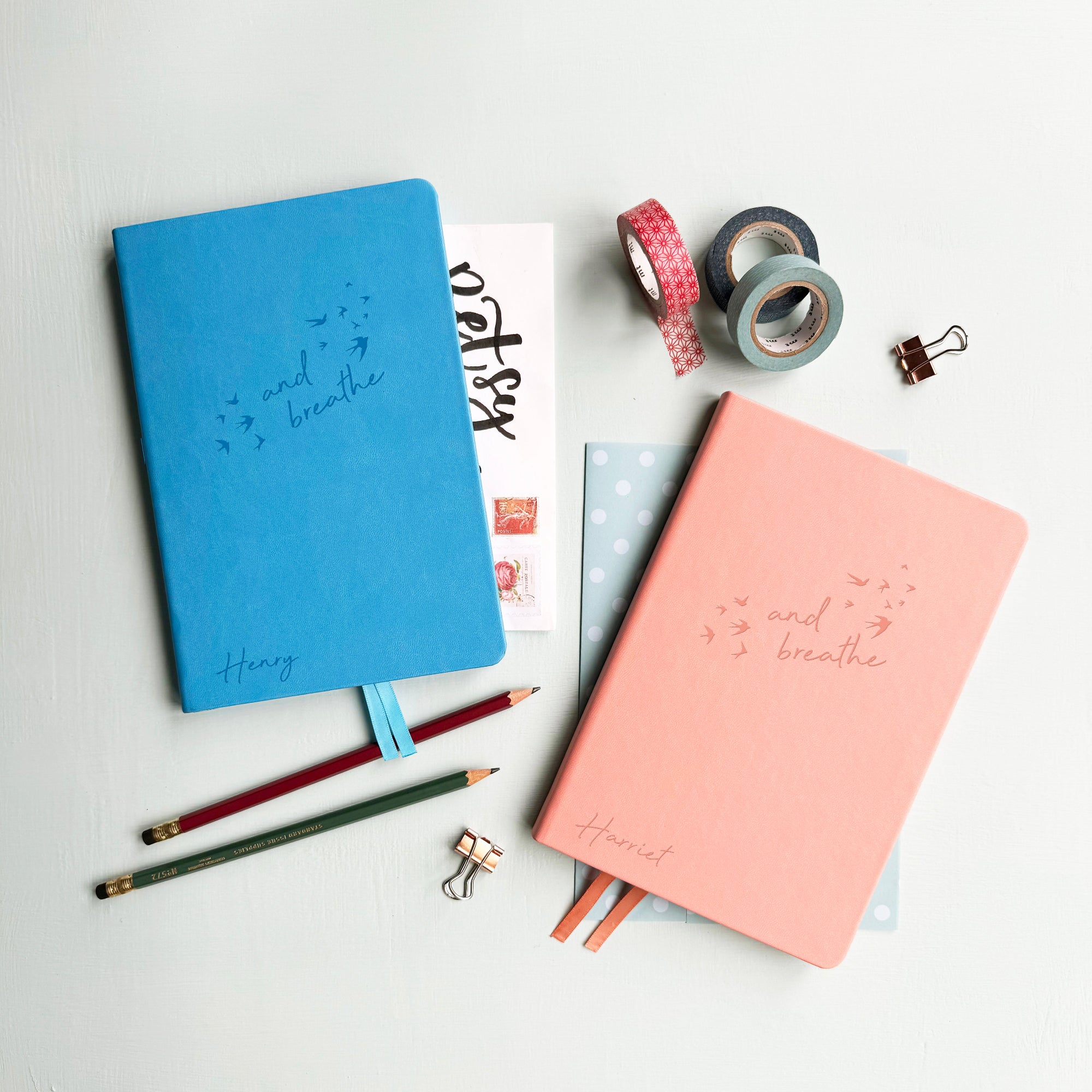 A top down image of two vegan leather notebooks, one blue and one pink. They are resting on a pale aqua table. The front of the notebook features the words "and breathe" surrounded by birds. The name Henry is engraved on the blue notebook and Harriet is engraved on the pink journal.  Surrounding the book are various stationary items.