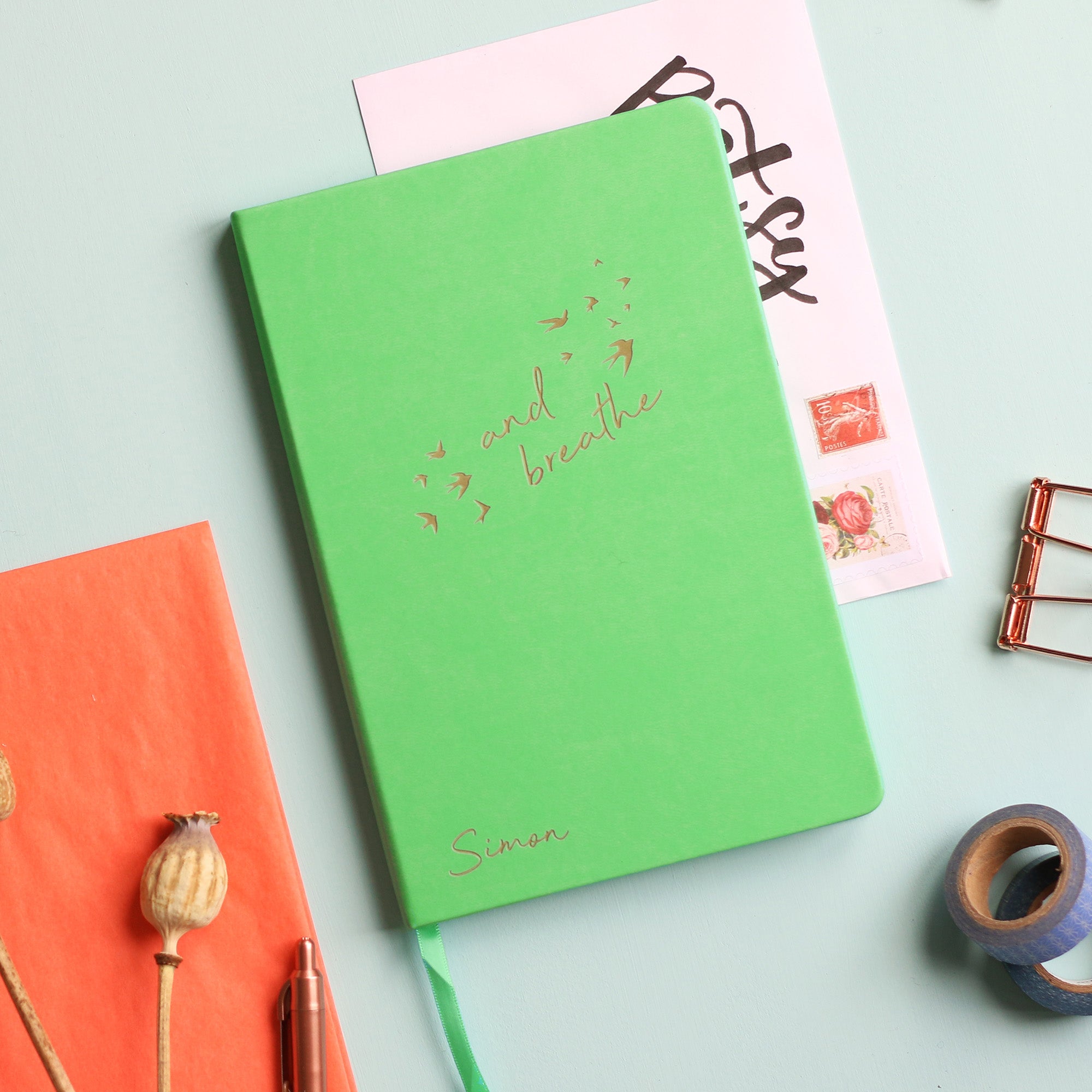 A top down image of a green vegan leather notebook resting on a pale aqua table. The front of the notebook features the words "and breathe" surrounded by birds. The personalised engraving reads Simon. Surrounding the book are various stationary items and dried flowers.