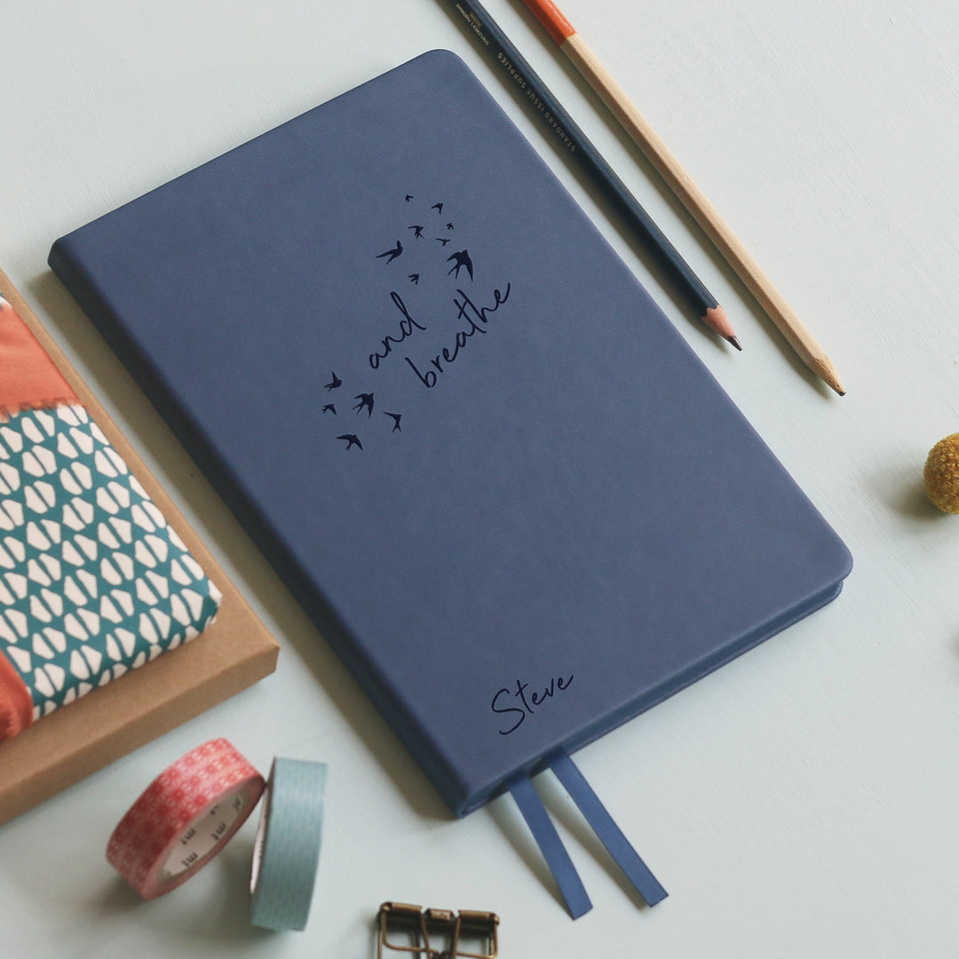 An image of a mid blue vegan leather notebook resting on a pale aqua table. The front of the notebook features the words and breathe surrounded by birds. The personalised engraving reads Steve. Surrounding the book are various stationary items.