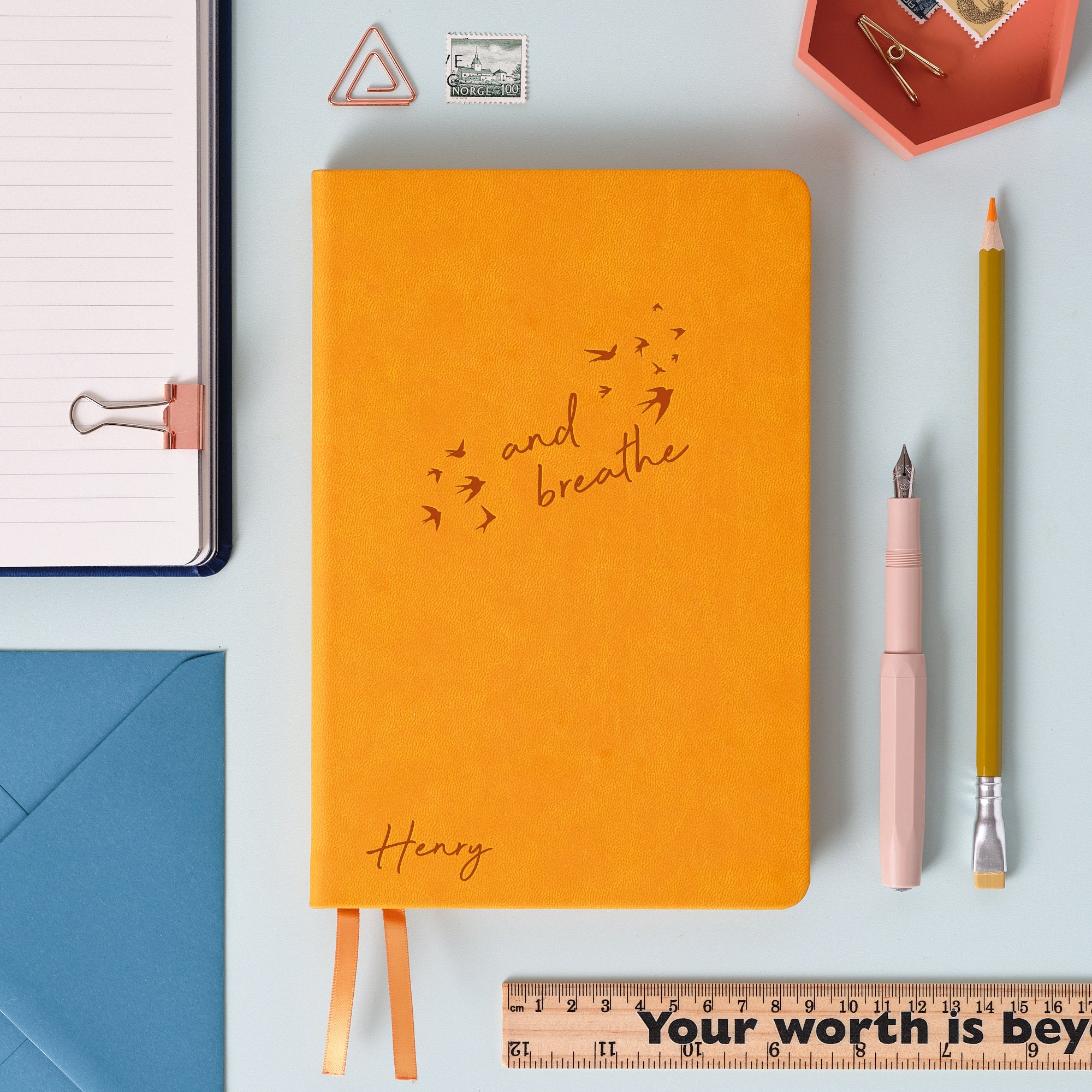 A top down image of a Mustard Yellow vegan leather notebook resting on a pale aqua table. The front of the notebook features the words "and breathe" surrounded by birds. The personalised engraving reads Henry. Surrounding the book are various stationary items and dried flowers.