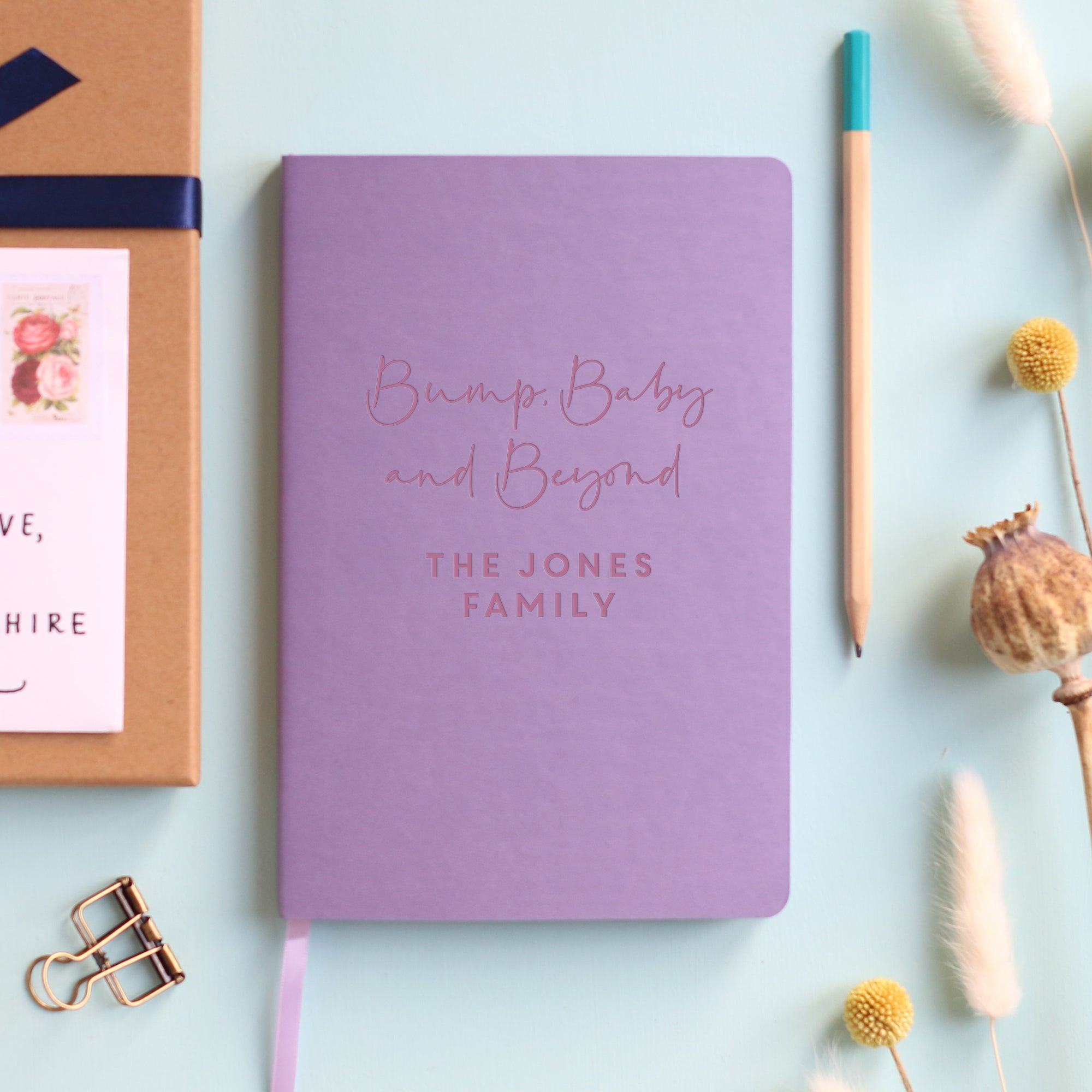 A top down image of a pink vegan leather notebook resting on a pale aqua table. The front of the notebook features the phrase Bump, Baby and Beyond. The personalised engraving reads Alice & Joe. Surrounding the book are various stationary items and dried flowers.