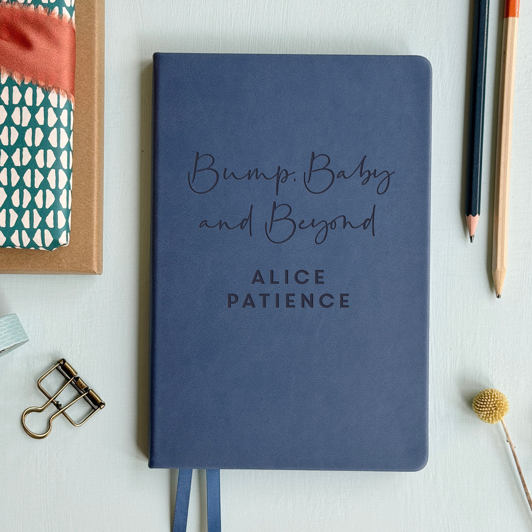 A top down image of a mid blue vegan leather notebook resting on a pale aqua table. The front of the notebook features the words Bump, Baby and Beyond. The personalised engraving reads Alice Patience. Surrounding the book are various stationary items and dried flowers.