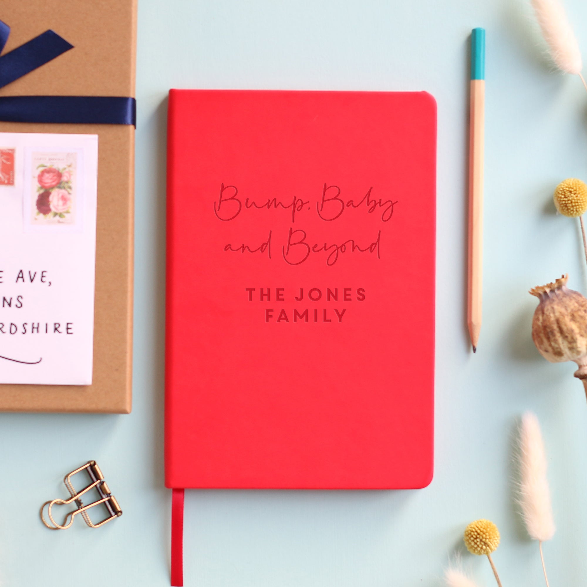 A top down image of a red vegan leather notebook resting on a pale aqua table. The front of the notebook features the phrase Bump, Baby and Beyond. The personalised engraving reads The Jones Family. Surrounding the book are various stationary items and dried flowers.