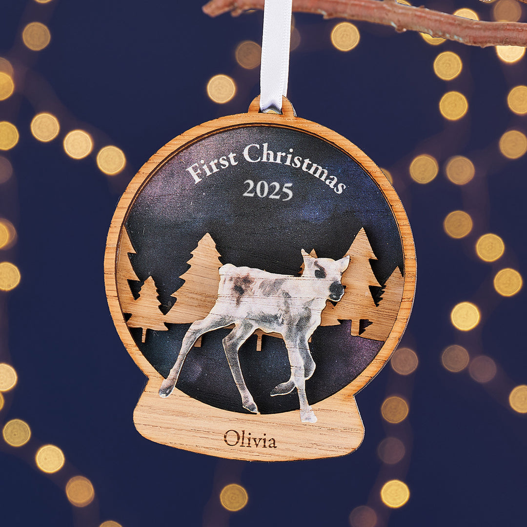 Wooden Christmas ornament with a reindeer, 'First Christmas 2025', and 'Olivia' on a dark background with lights.
