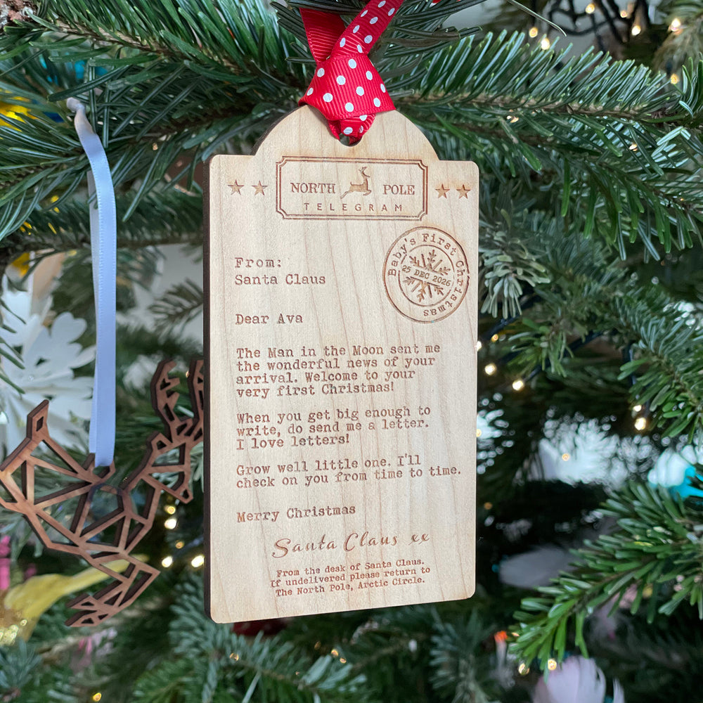 A wooden Baby's first Christmas ornament tag with a red spotty ribbon, engraved with a telegram from Santa Claus, displayed on a Christmas tree.