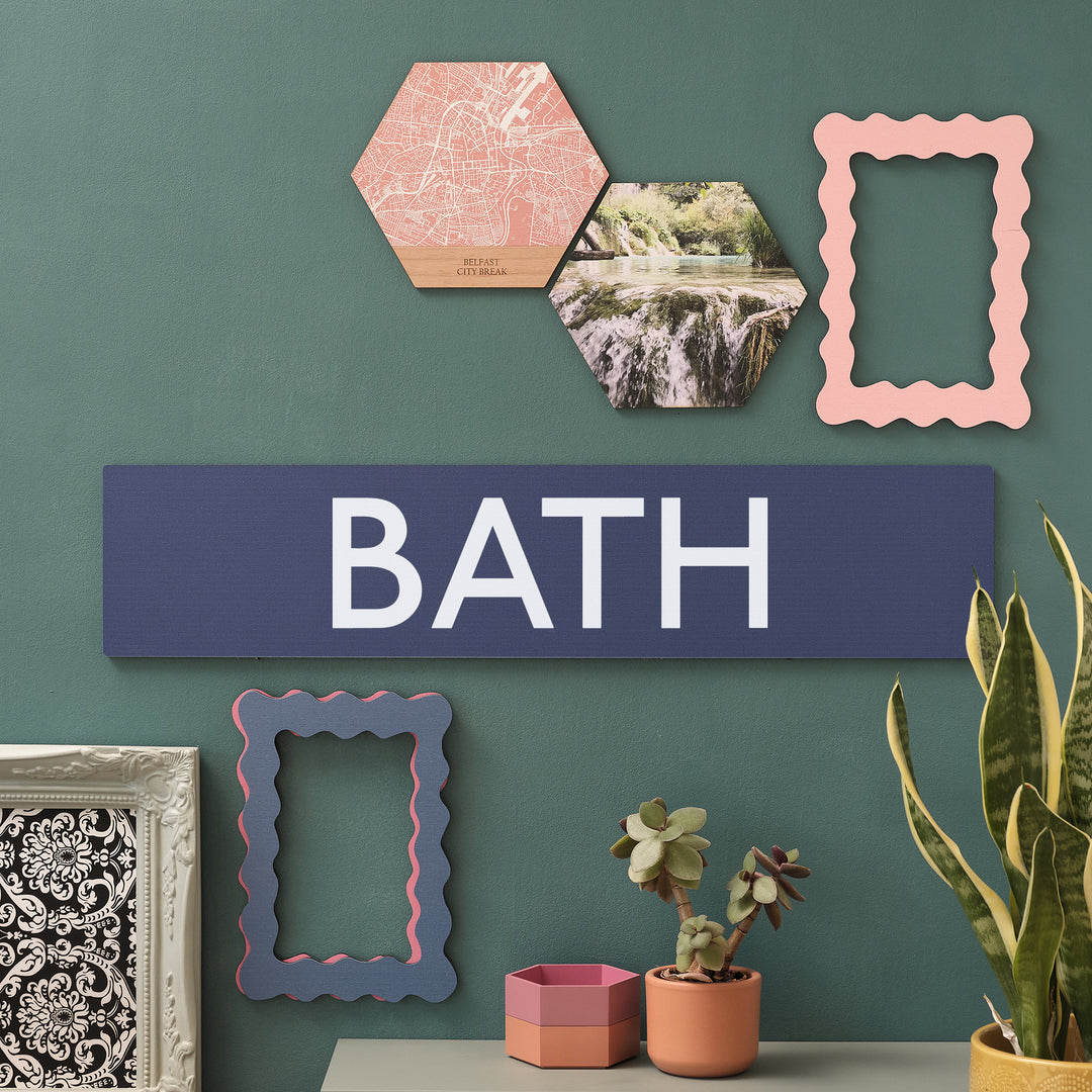 Large size dark blue wooden wall art print for maximalist travel lovers. Large white text that says 'BATH' for UK city break commemorative display hung on a green wall with a variety of frames and plants around.