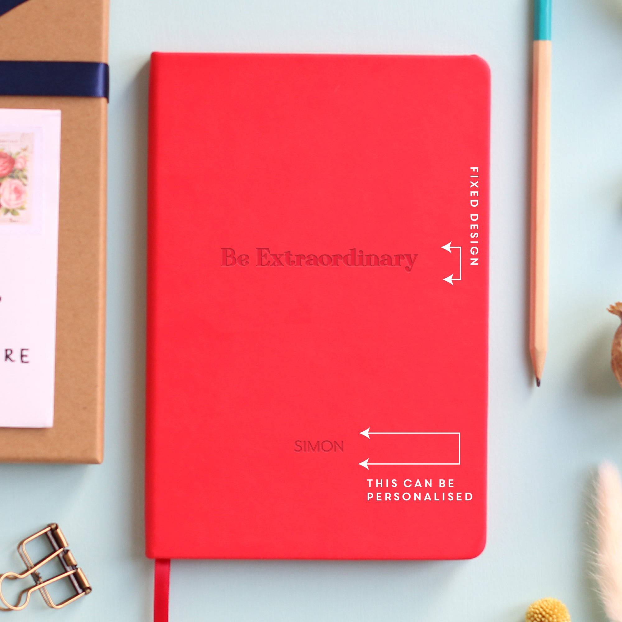 A top down image of a red vegan leather notebook resting on a pale aqua table. The front of the notebook features the words Be Extraordinary. The personalised engraving reads Simon. There is additional text showing the areas that can be customised. Surrounding the book are various stationary items and dried flowers.