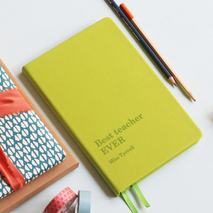 Green notebook with 'Best teacher EVER' text, surrounded by colorful stationery items on a white background