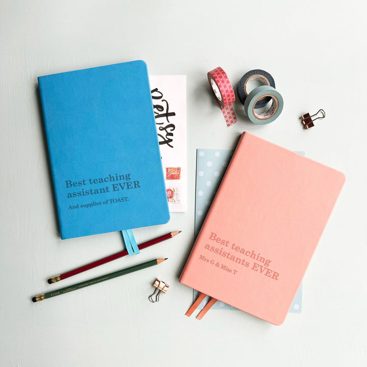 Two notebooks, blue and pink teaching assistant gift notebook engraved best teaching assistant ever text, atop pale grey background with assorted stationary surrounding.